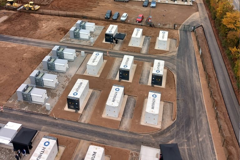 Luftbild eines Batteriegroßspeichers: weiße Container in Reihen auf einer eingezäunten, planierten Fläche, rechts eine Landstrasse, links ein Acker.